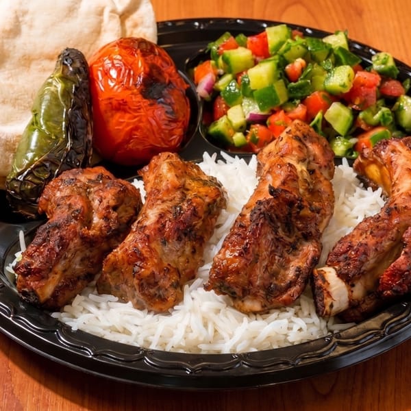 Grilled chicken ribs, rice, salad, pita bread, and roasted vegetables on a black plate.