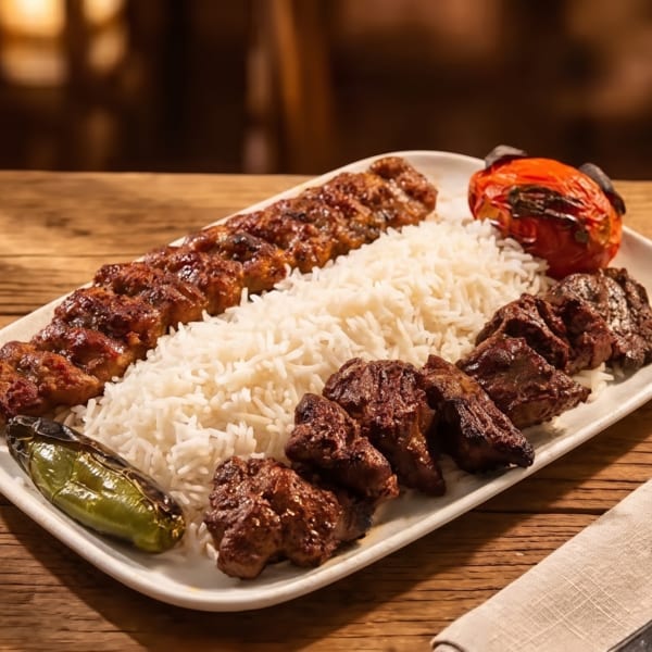 A white platter of Persian kabob, rice, grilled vegetables, and a glass of tea on a wooden table.